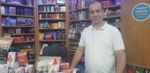 Photo of a smiling man in a corner shop, with watches, e cigarattes and child’s sweets behind him.