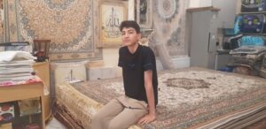 a teenager, sat on a stack of persian rugs, looking quizzically to the camera.