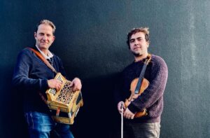 James Delarre and Saul Rose stand against dark wall holding melodean and fiddle