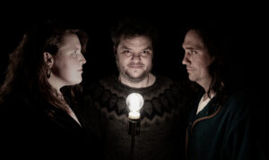 Ward, Knutur & Townes in a dark black background stand around facing a small lit lightbulb