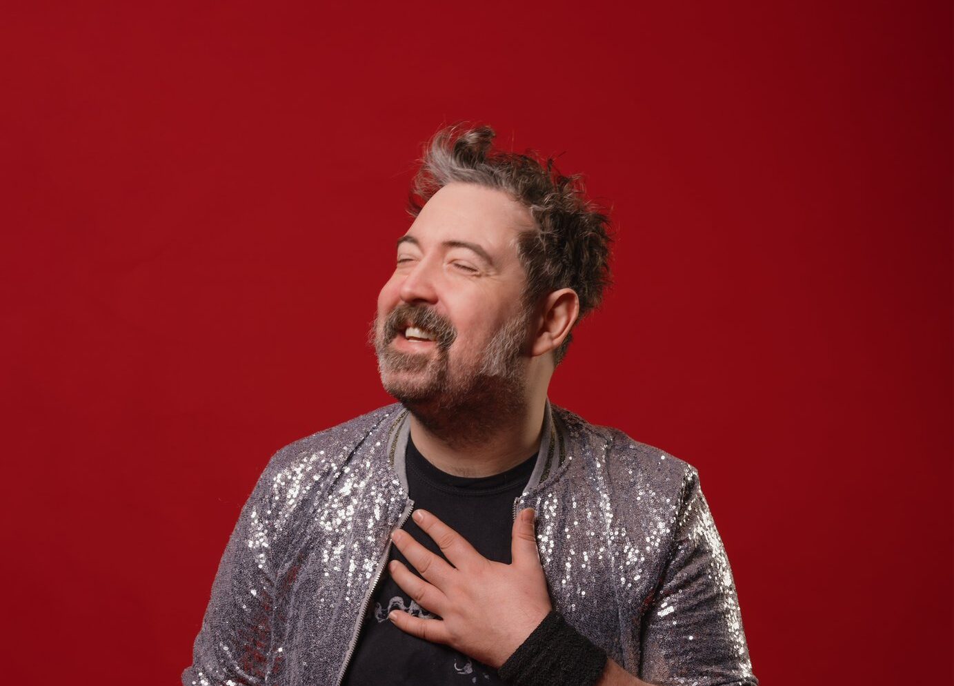 nick helm against a red background wearing a black t shirt and silver jacket
